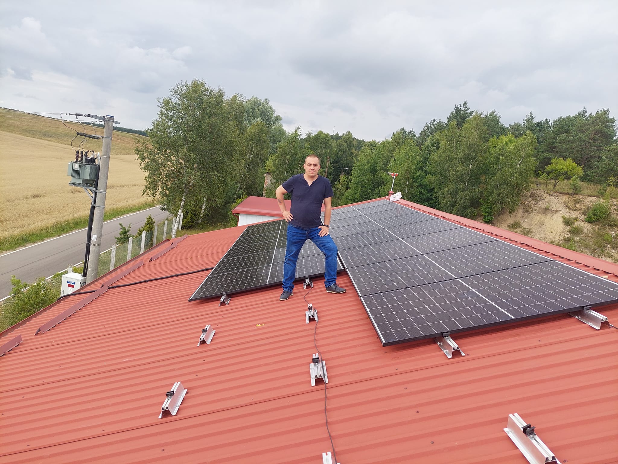 Montaż paneli fotowoltaicznych na czerwonym dachu z blachy trapezowej. Mężczyzna w jeansach stoi na dachu obok zamontowanych paneli. W tle drzewa i słup elektryczny.