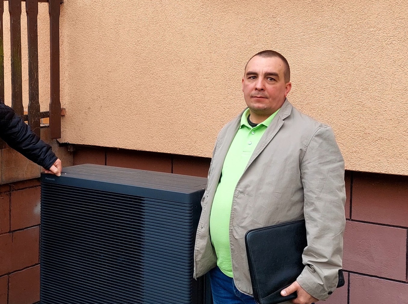 Mężczyzna w jasnej marynarce stoi obok pompy ciepła Panasonic, trzymając czarną teczkę. Część dłoni widoczna na pompie. Tło: beżowa ściana i fragment ceglanego muru.