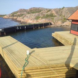 DIGAB - Nowy drewniany taras nad wodą, połączony z pomostem w zatoce, widok na skaliste wybrzeże i czerwoną szopę, cień fotografa na tarasie.