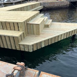 DIGAB - Nowy drewniany pomost z schodami, zbudowany nad wodą, z widocznymi elementami rustykalnego nabrzeża w tle i żółtym budynkiem na brzegu.