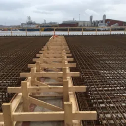 Siatka zbrojeniowa przygotowana do zalania betonem, z drewnianym pomostem roboczym, w tle widok na port z budynkami przemysłowymi i statkiem.