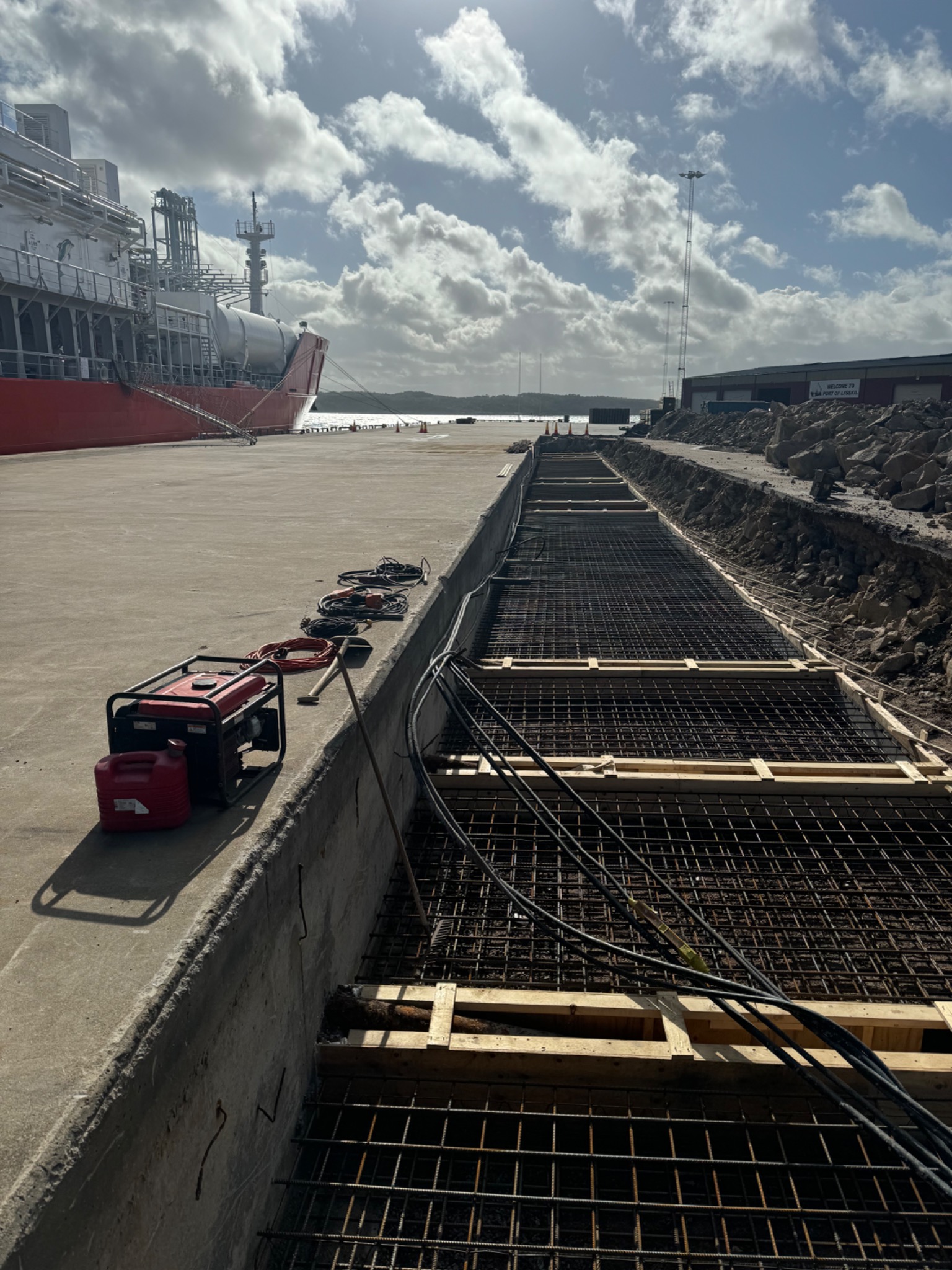 Zbrojenie fundamentów pod budowę nabrzeża portowego, widoczny generator prądu, narzędzia i zacumowany statek w tle.