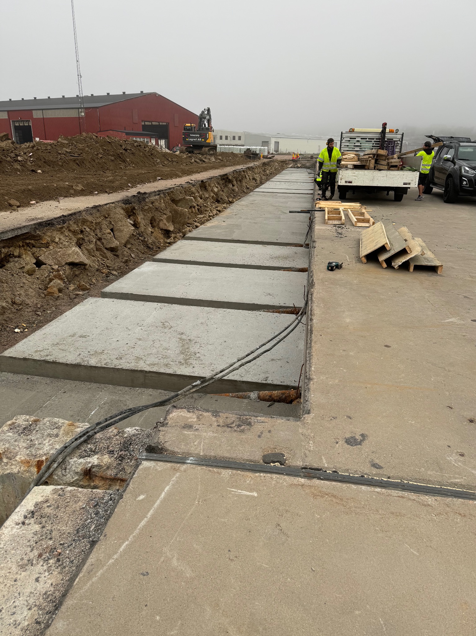 Betonowe płyty ułożone w wykopie, widoczni pracownicy w odblaskowych kamizelkach i ciężarówka z drewnianymi elementami, w tle budynek przemysłowy i koparka.