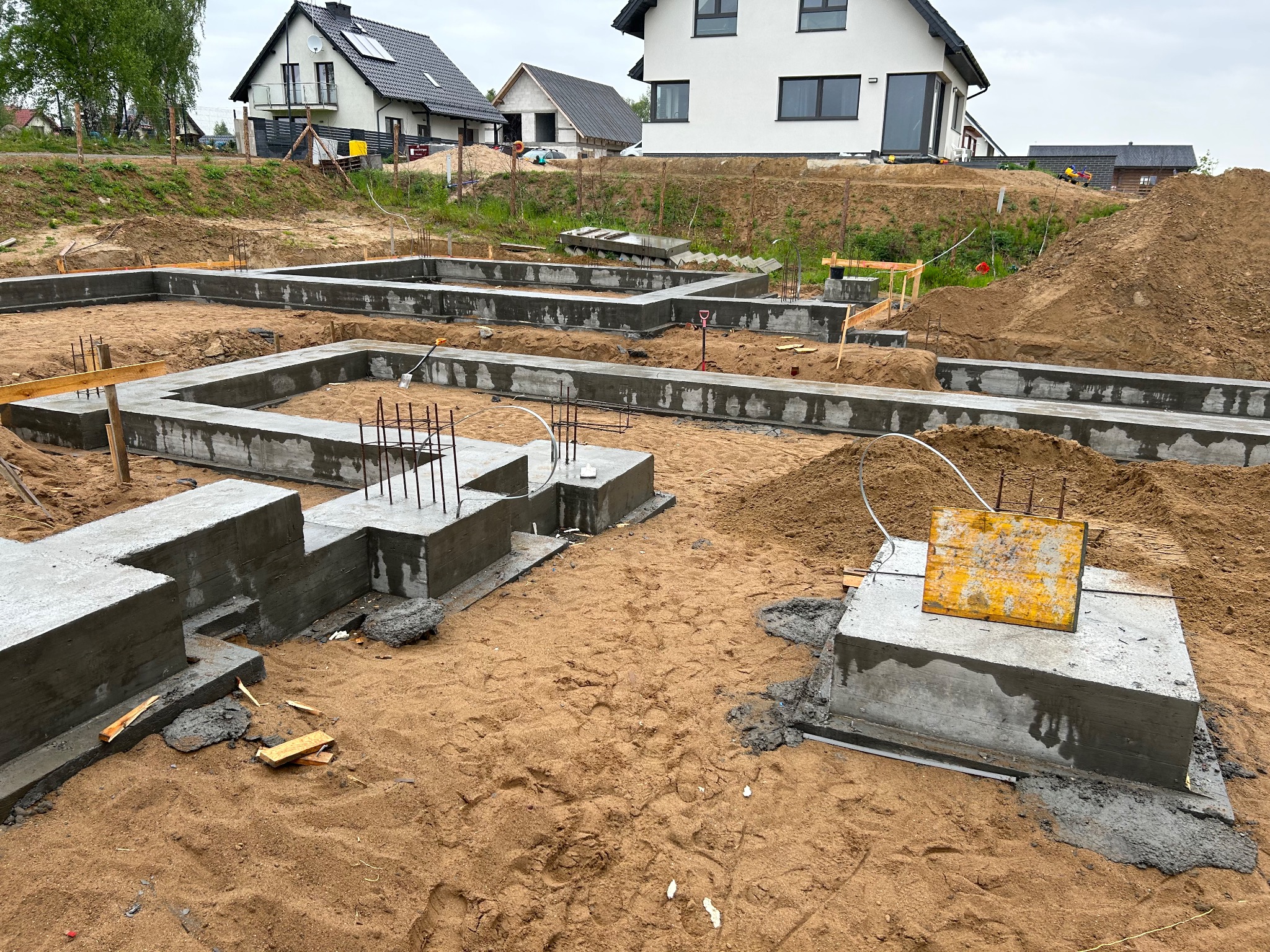 Wylane fundamenty pod budynek mieszkalny, widoczne zbrojenia, szalunki i świeży beton, w tle domy jednorodzinne.