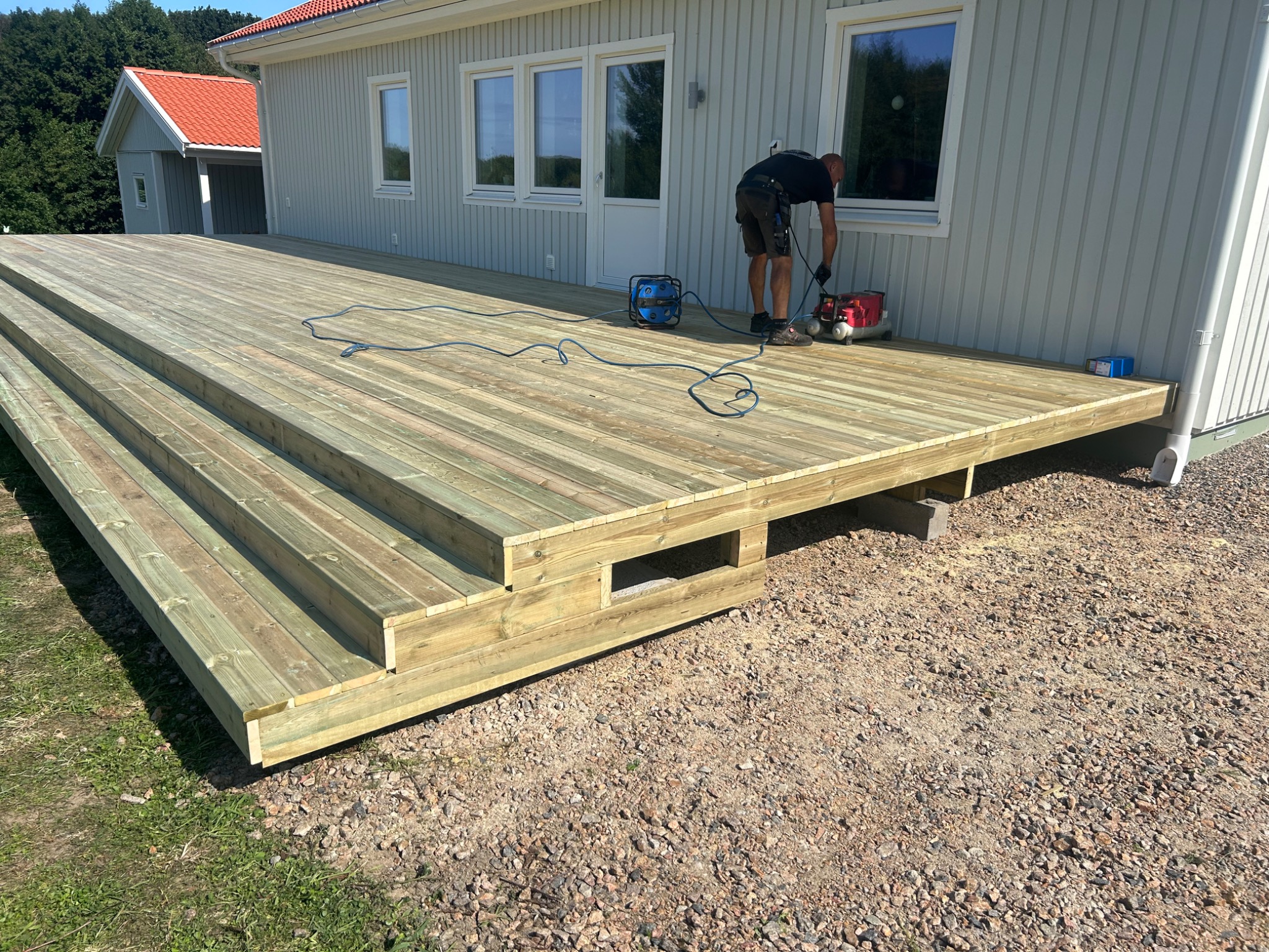 Nowo wybudowany, surowy taras z drewna z trzema stopniami, z pracownikiem szlifującym powierzchnię przy użyciu szlifierki elektrycznej i odkurzacza przemysłowego, na tle domu z szarą elewacją.