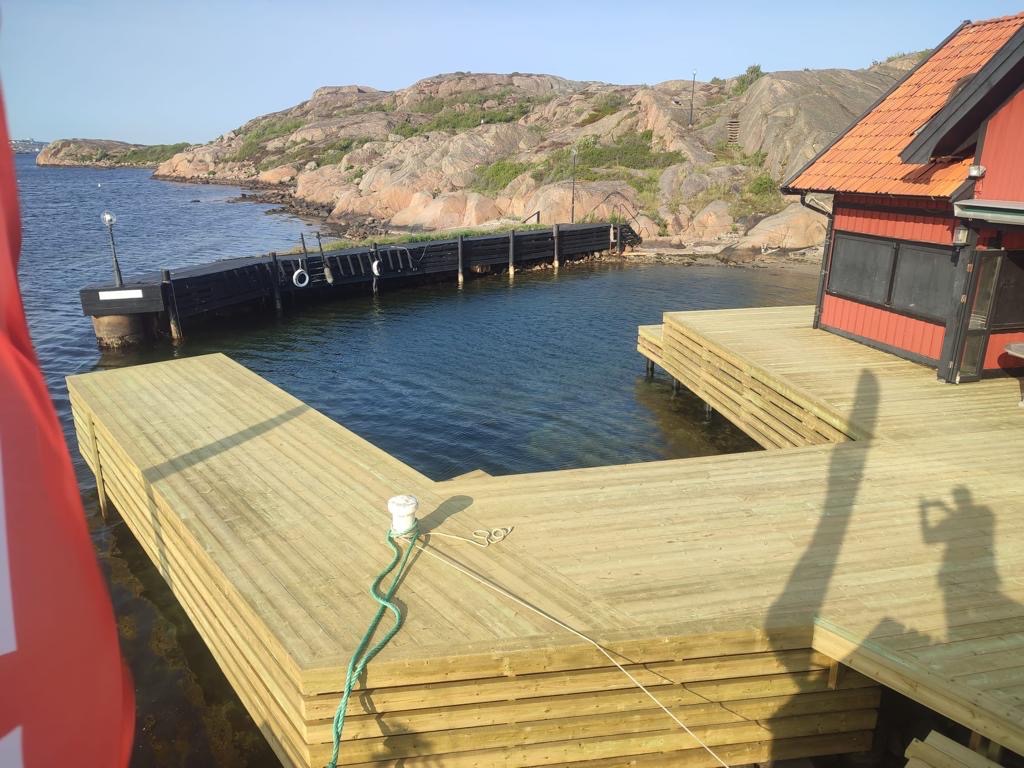 Nowy drewniany taras nad wodą, połączony z pomostem w zatoce, widok na skaliste wybrzeże i czerwoną szopę, cień fotografa na tarasie.