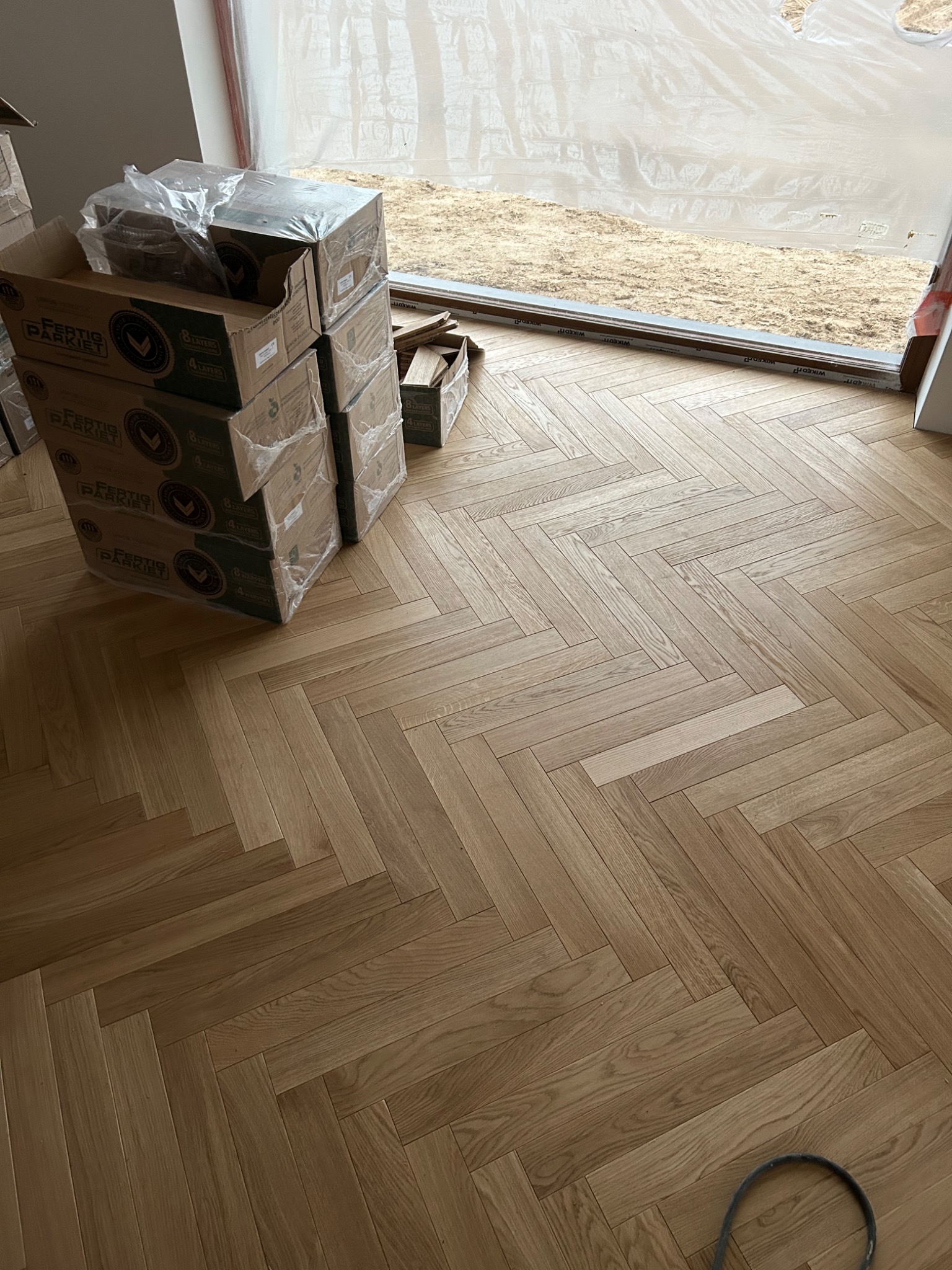Ułożona podłoga z parkietu w jodełkę, z kartonami parkietu Fertig Parkiet w tle i widokiem na zewnątrz przez okno zabezpieczone folią malarską.