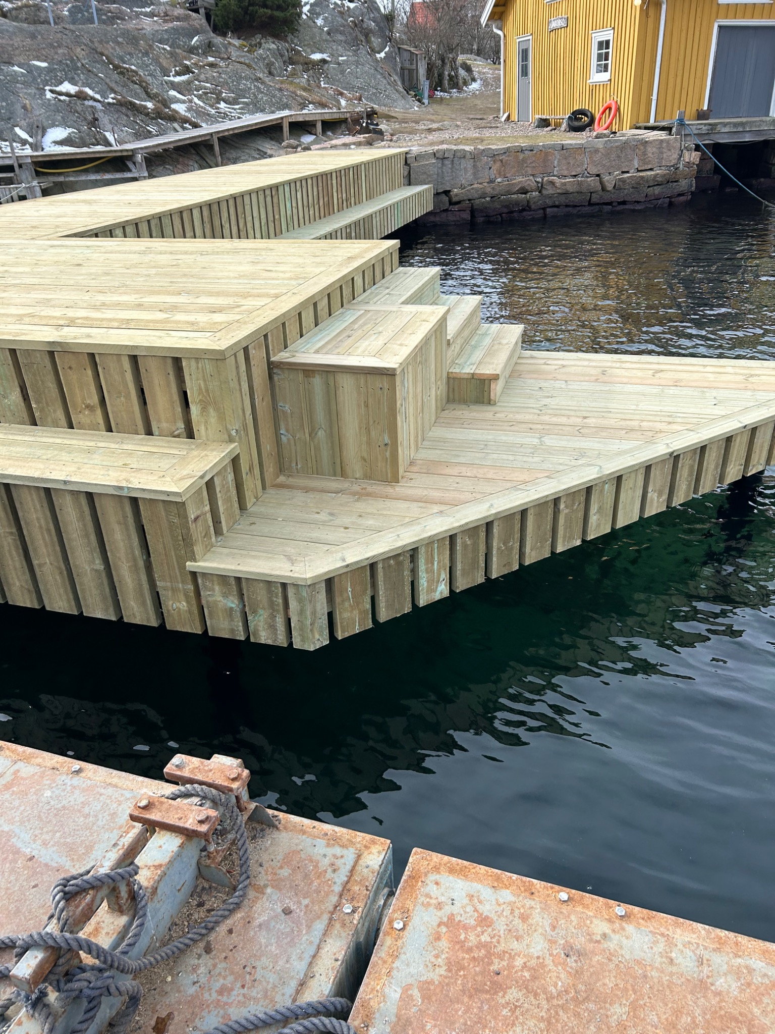 Nowy drewniany pomost z schodami, zbudowany nad wodą, z widocznymi elementami rustykalnego nabrzeża w tle i żółtym budynkiem na brzegu.