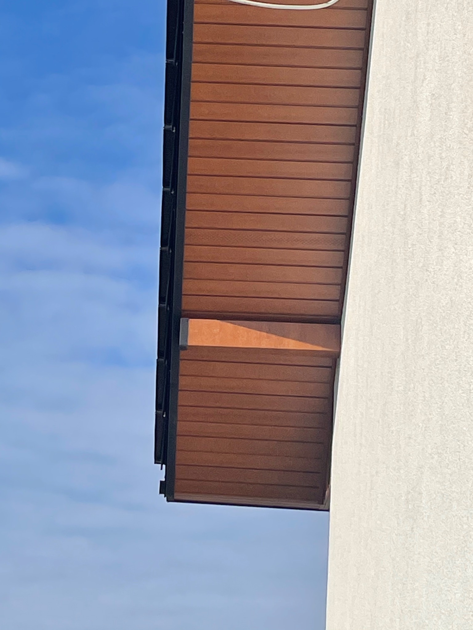 Fragment elewacji budynku z brązową podbitką dachową z desek kompozytowych i białą ścianą zewnętrzną na tle błękitnego nieba.