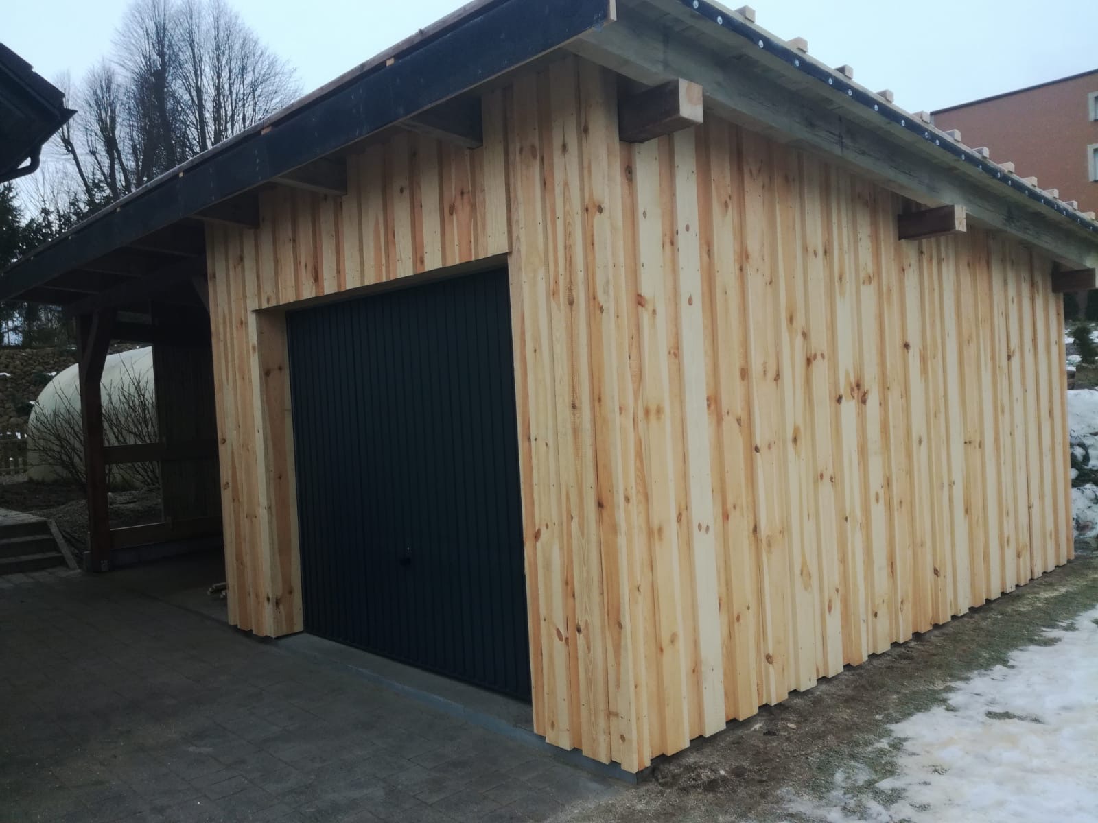 Nowo wybudowany garaż z pionową boazerią z jasnego drewna i czarną bramą segmentową, widoczny fragment zadaszenia i resztki śniegu na ziemi.