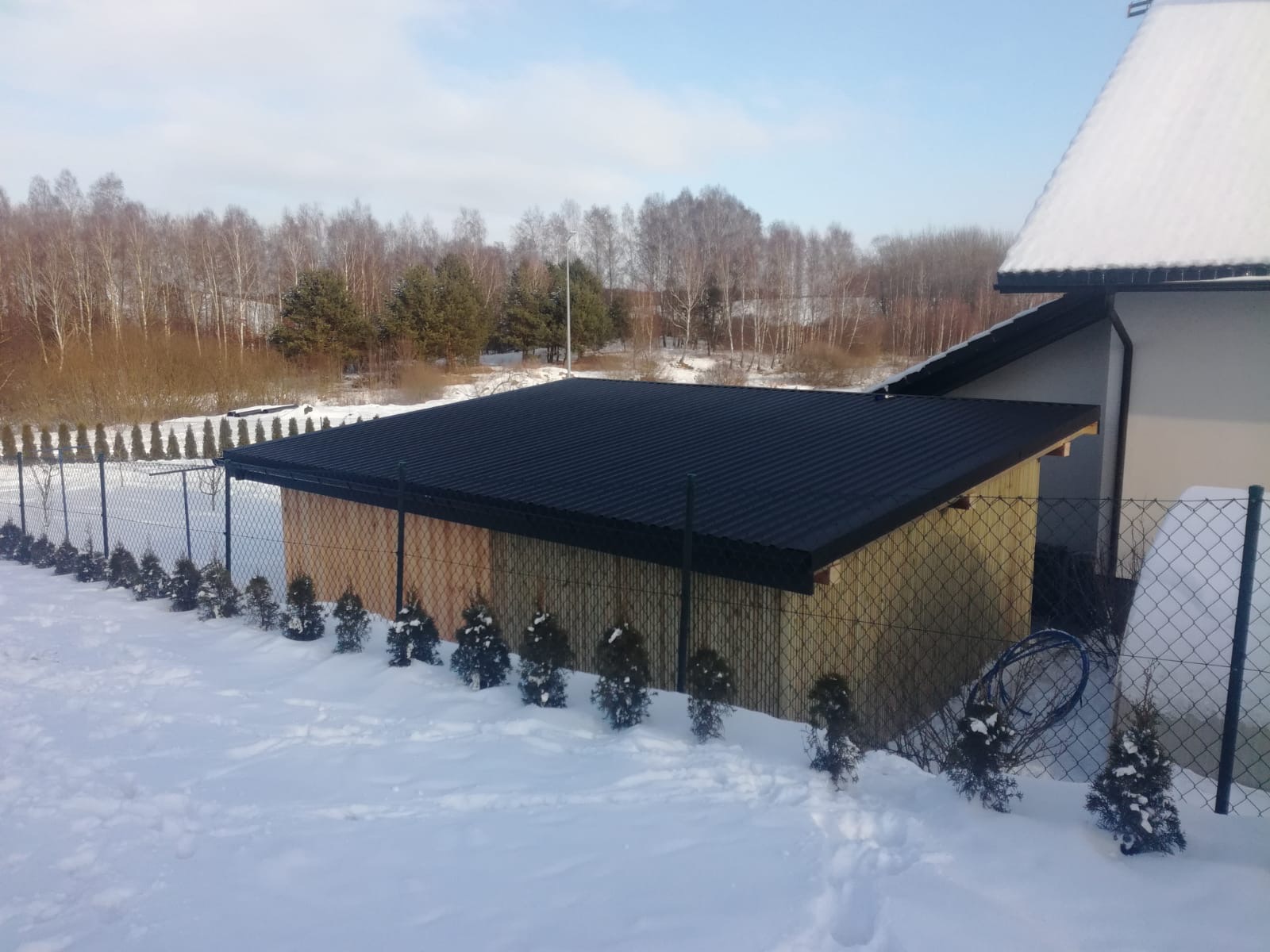 Zadaszenie tarasu z ciemną, falistą blachą, przylegające do budynku, otoczone śniegiem, siatką ogrodzeniową i rzędem małych iglaków.