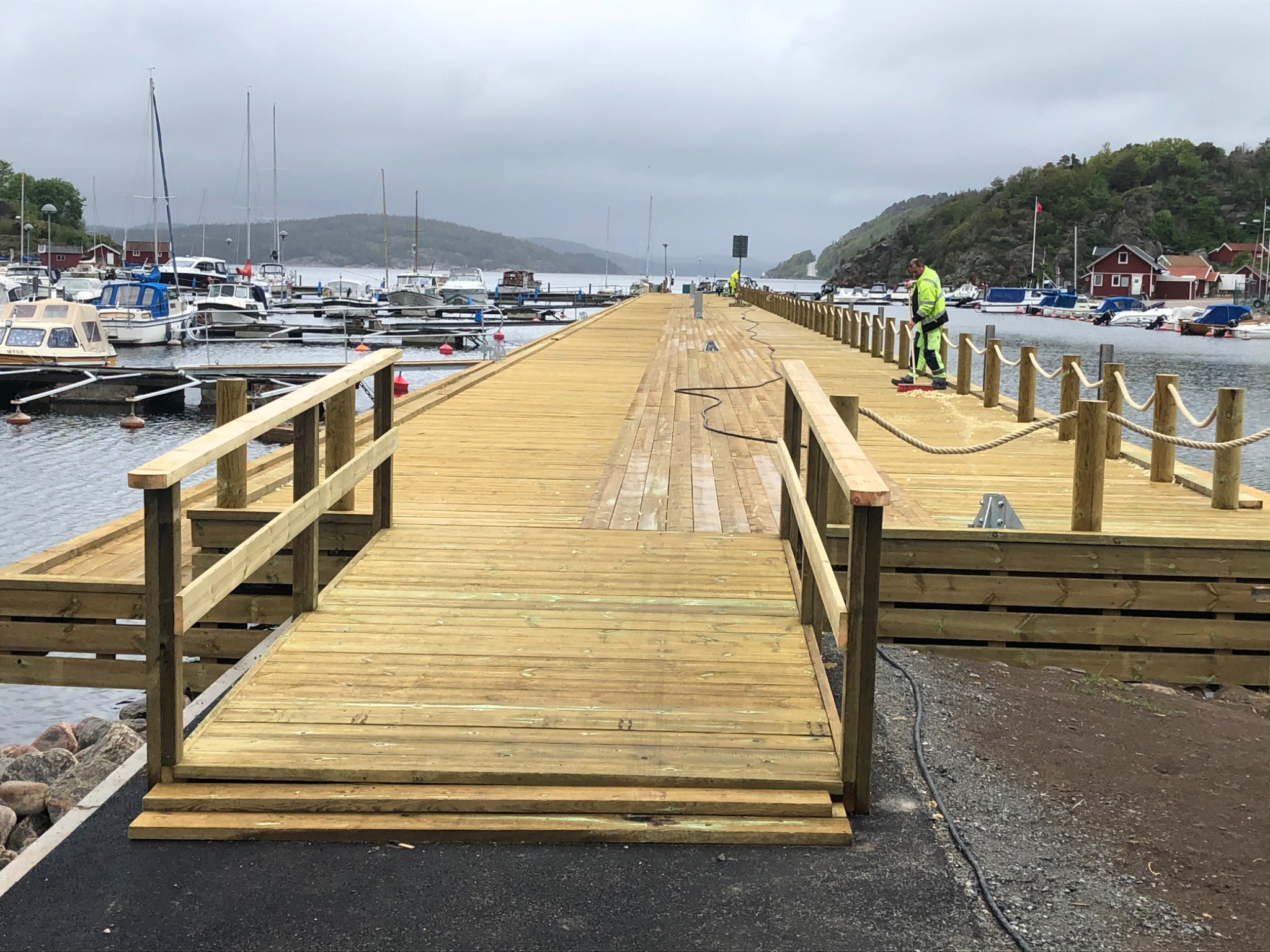 Nowy, długi, drewniany pomost w marinie z łodziami, z pracownikiem w odblaskowej kamizelce zamiatającym powierzchnię, widok na góry i pochmurne niebo w tle.