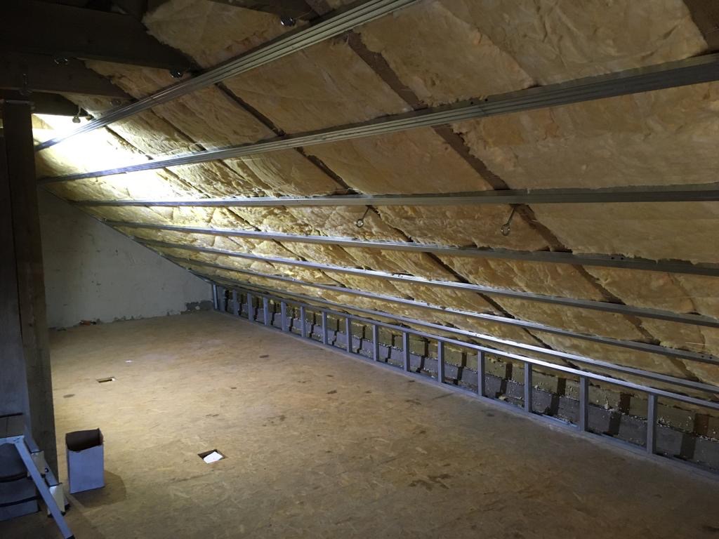 Poddasze w trakcie ocieplania wełną mineralną, widoczne metalowe profile mocujące, surowe wykończenie podłogi z płyty OSB, częściowo oświetlone naturalnym światłem z okna dachowego.