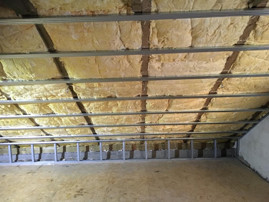 Poddasze w trakcie izolacji, widoczne metalowe profile konstrukcyjne i wełna mineralna między nimi, surowa podłoga z płyt OSB, fragment ściany w tle.