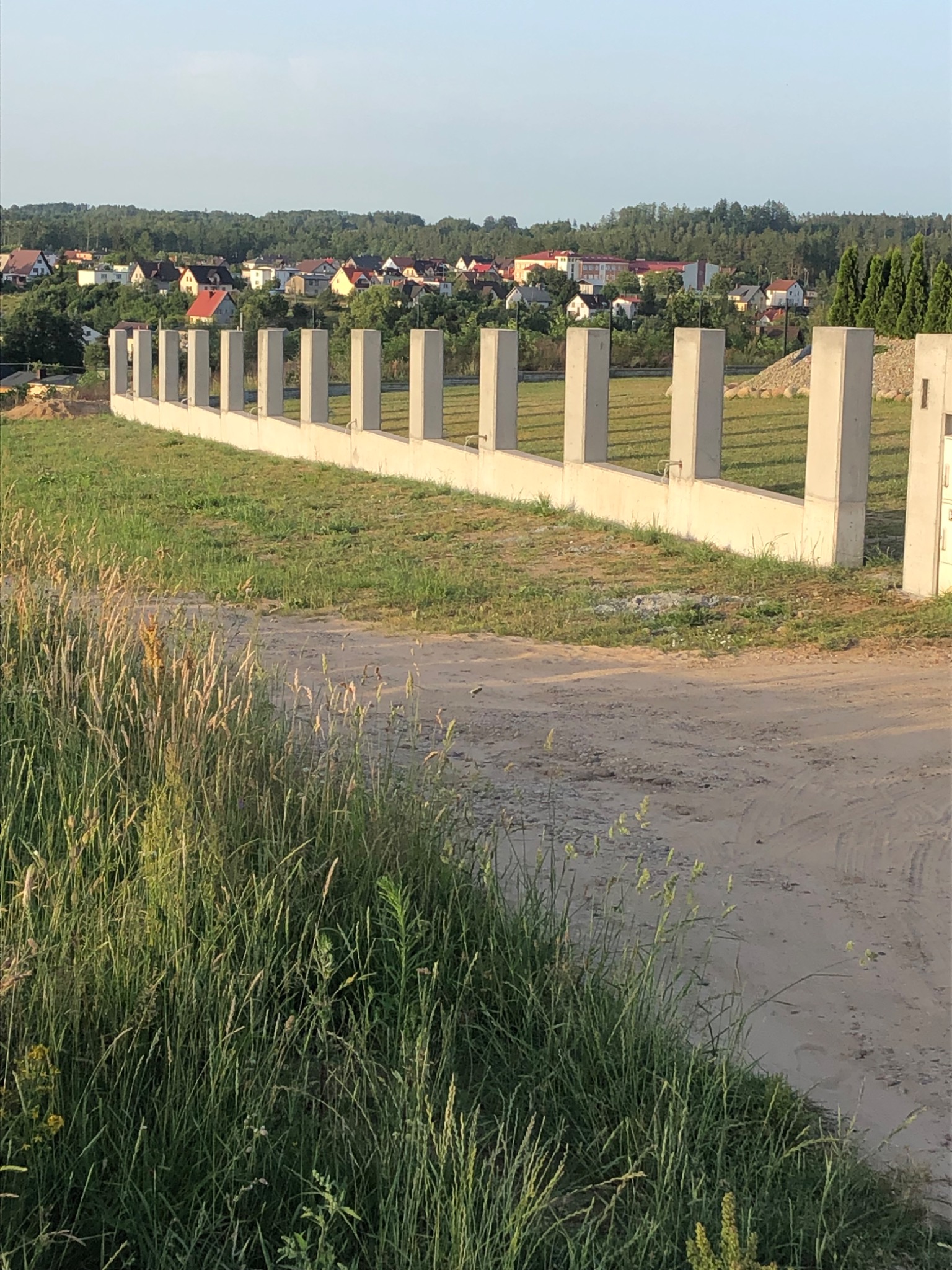 Wznoszenie betonowego ogrodzenia modułowego na nierównym terenie z widokiem na zabudowę jednorodzinną.