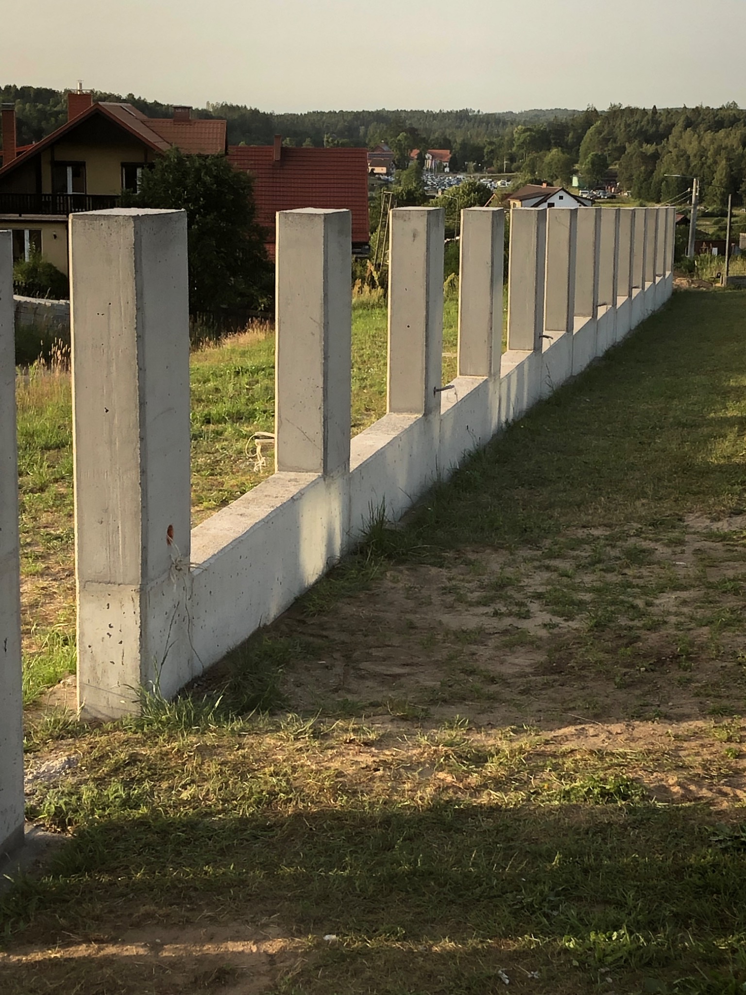 Surowy betonowy fundament ogrodzenia z wystającymi słupkami, widok z boku na nierówny teren z trawą i ziemią, w tle domy i drzewa w letnim świetle.