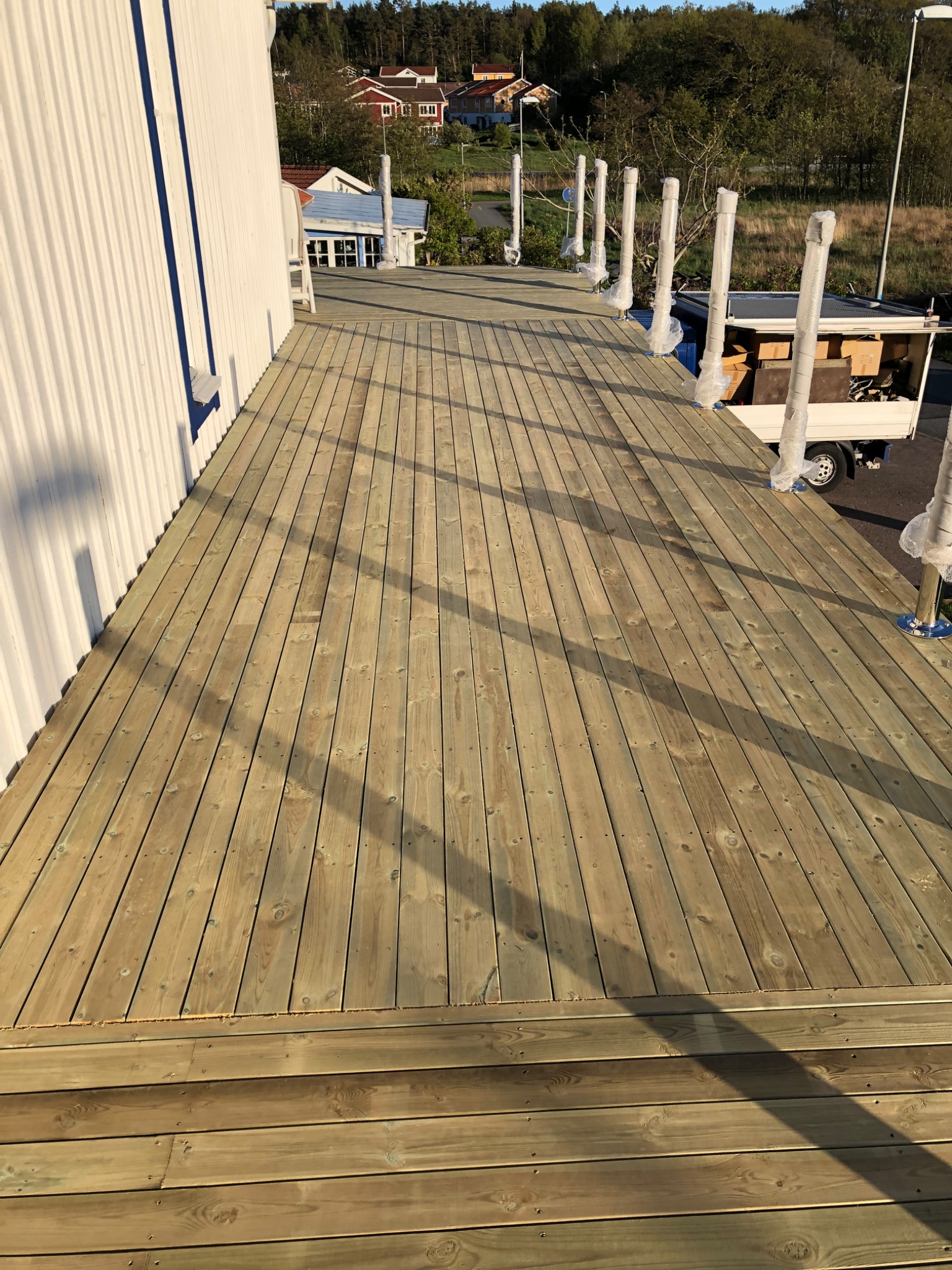 Nowy taras z desek w odcieniach zieleni, przylegający do budynku z białymi panelami, z widocznymi elementami konstrukcyjnymi zabezpieczonymi folią, a w tle zabudowa jednorodzinna i zieleń.