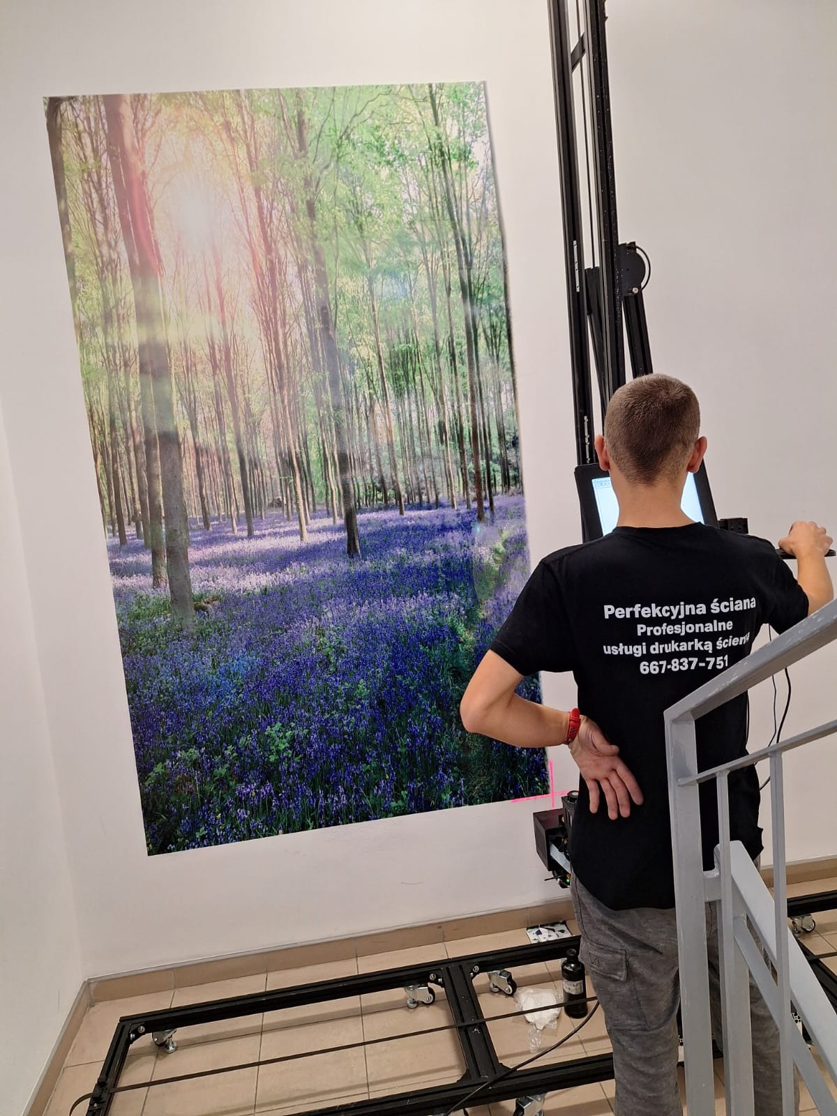 Mężczyzna obsługuje drukarkę ścienną, nanosząc motyw leśny z kwitnącymi dzwonkami na ścianę. Widoczny fragment schodów i podłogi z beżowymi płytkami.