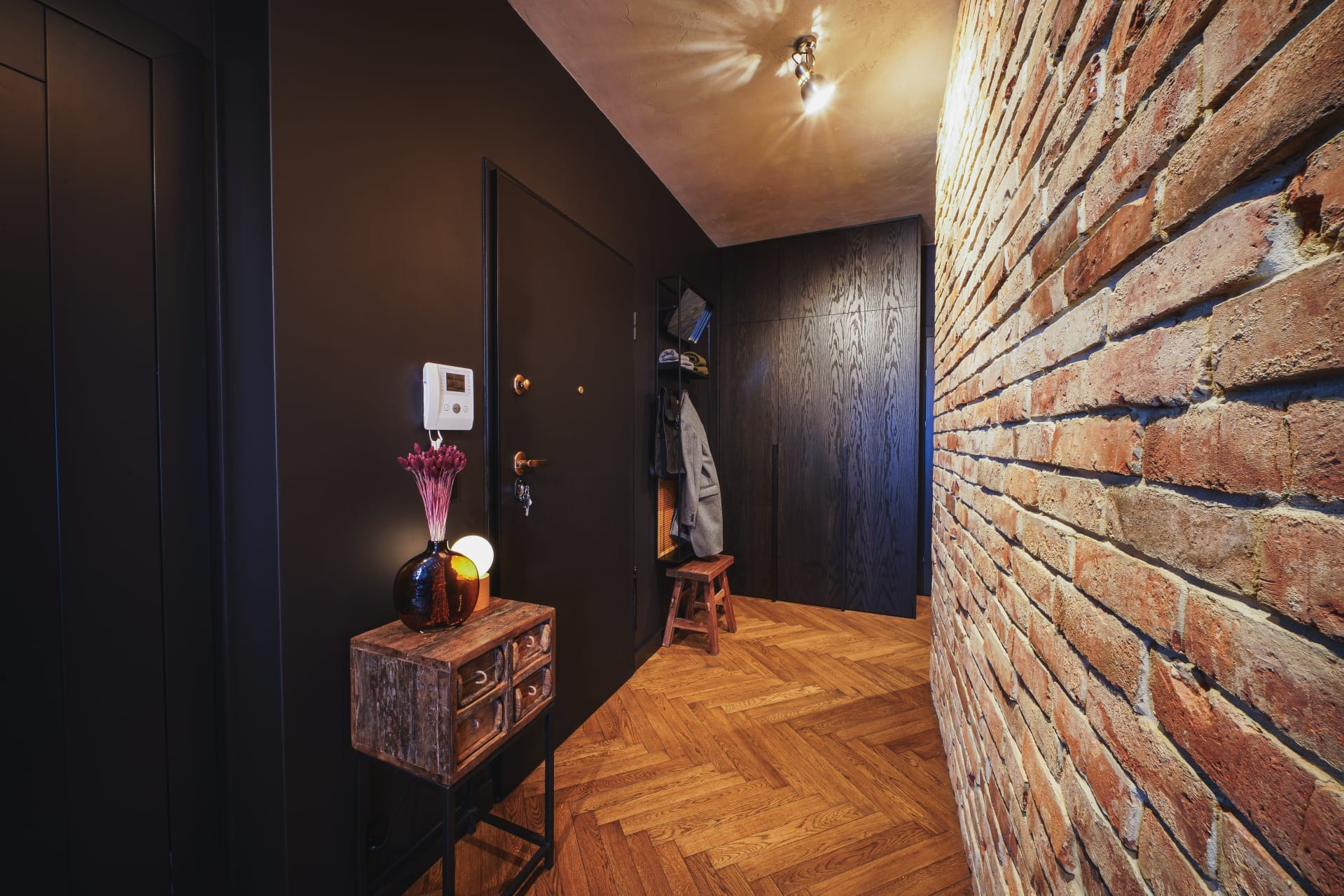 Nowoczesny przedpokój z ceglaną ścianą, parkietem w jodełkę, ciemnymi drzwiami i szafą w zabudowie. Na pierwszym planie rustykalna komoda z dekoracyjnym wazonem i suszonymi kwiatami.