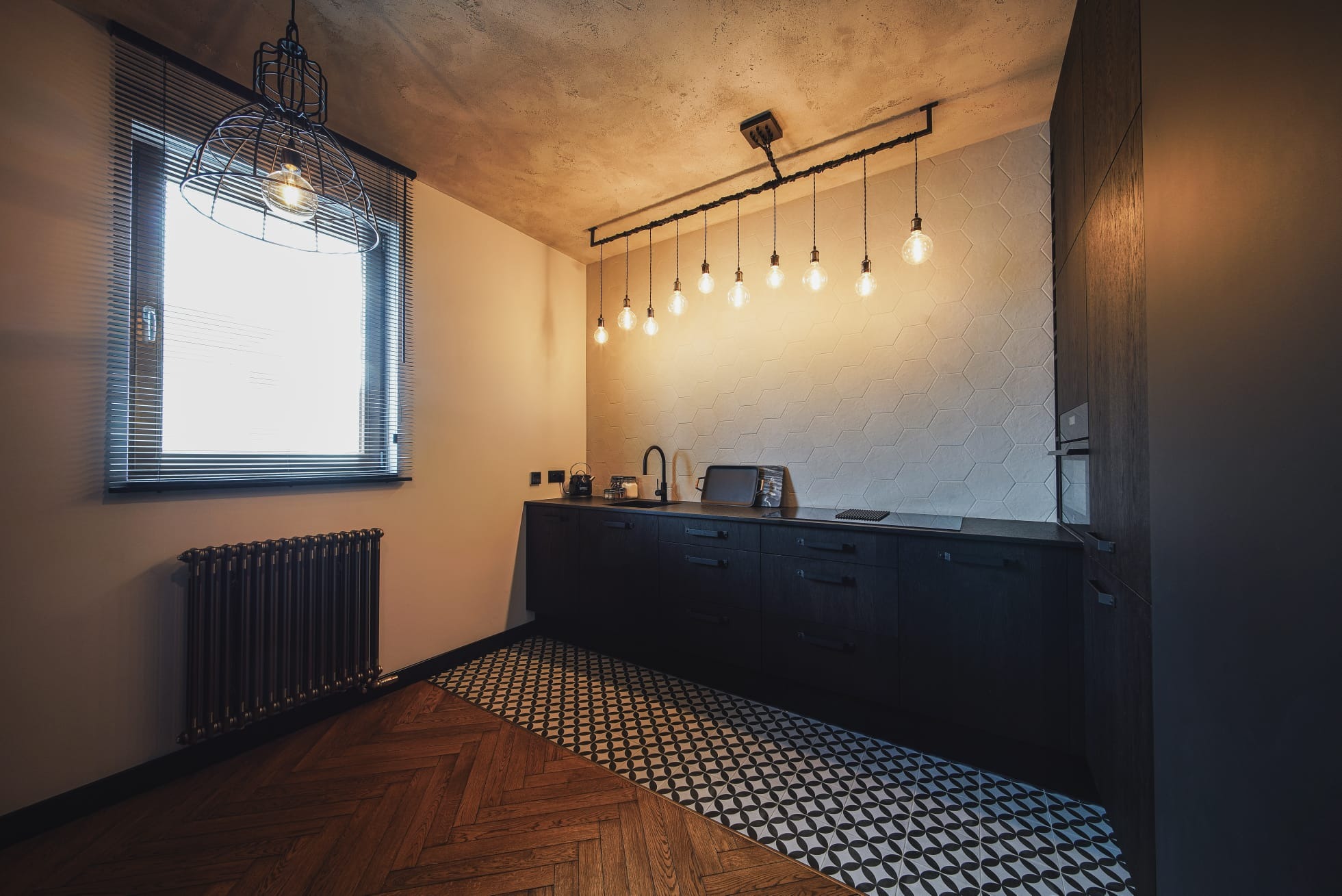 Nowoczesna kuchnia w Warszawie z czarnymi szafkami, heksagonalnymi płytkami ściennymi i designerskim oświetleniem, gdzie podłoga łączy parkiet w jodełkę z geometrycznym wzorem płytek.