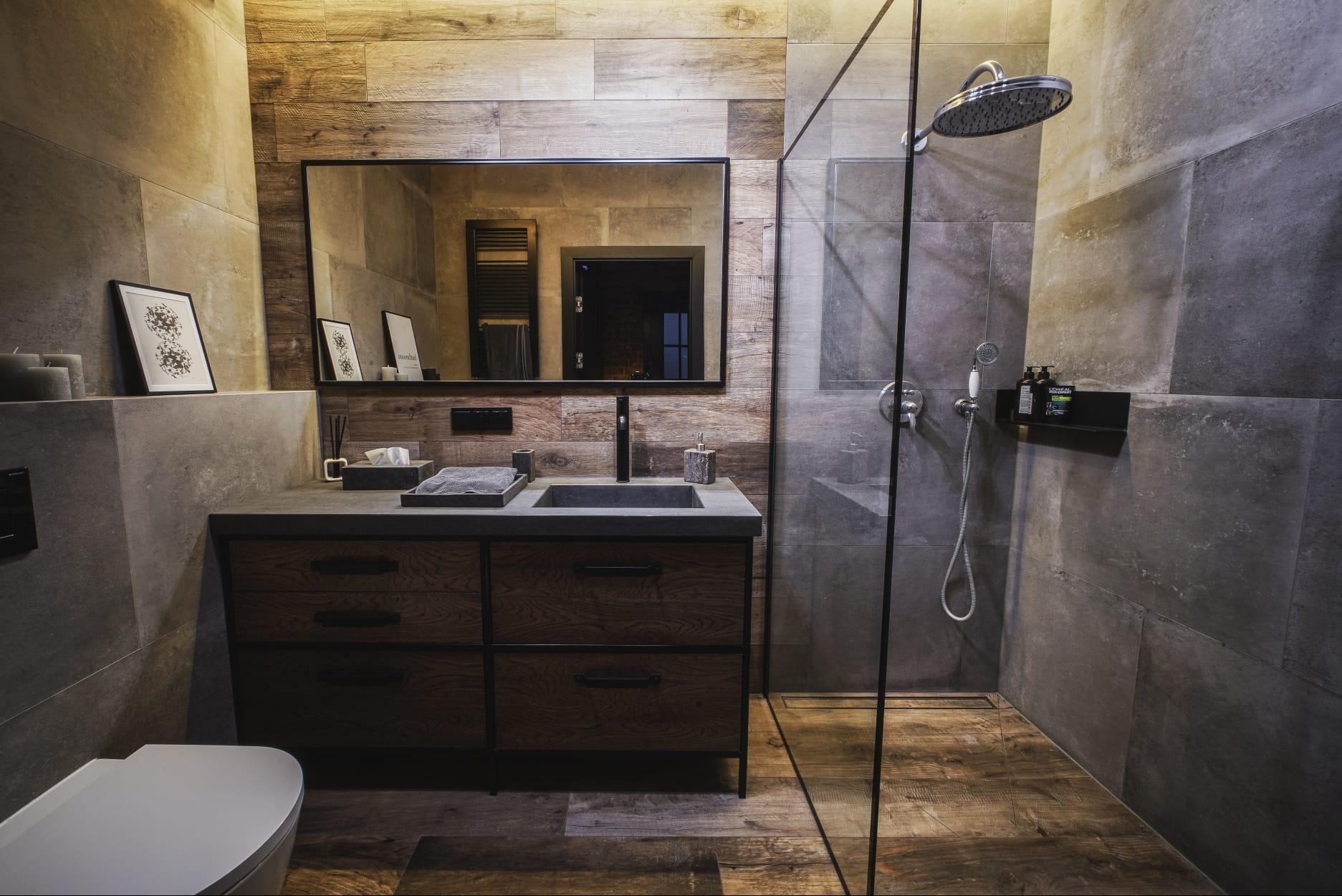 Nowoczesna łazienka z drewnianymi i szarymi płytkami, kabiną prysznicową typu walk-in i szafką pod umywalkę z ciemnego drewna z czarnymi okuciami, widok na lustro z odbiciem grzejnika.