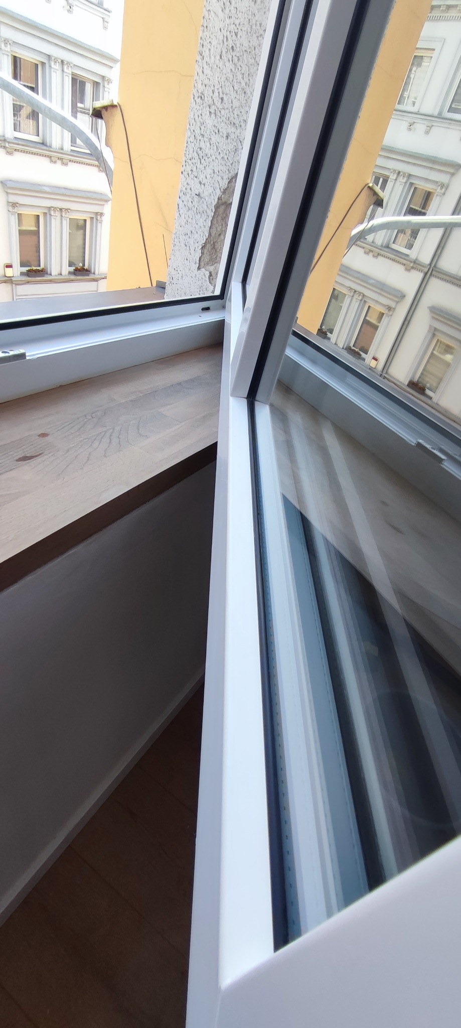 Otwarty biały profil okienny z widokiem na budynki odbijające się w szybie, z fragmentem drewnianego parapetu i szarej ściany z uszkodzonym tynkiem.