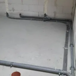 Szare rury instalacyjne prowadzone wzdłuż surowych ścian z betonu komórkowego, zabezpieczone pianką montażową w miejscach przejść, z widocznymi obejmami mocującymi i fragmentem podłogi z otworem.