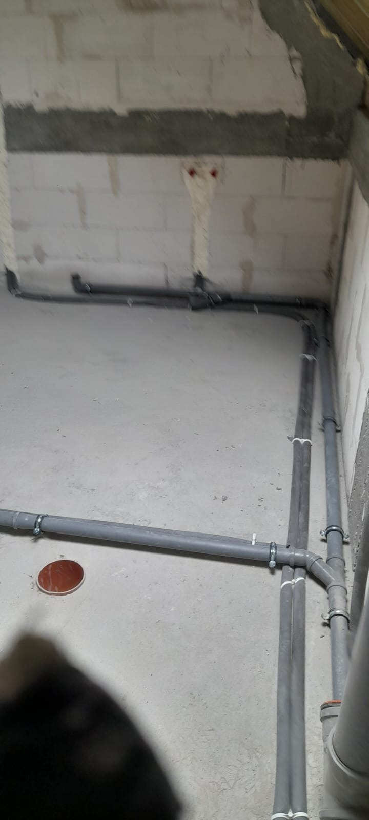 Szare rury instalacyjne prowadzone wzdłuż surowych ścian z betonu komórkowego i podłogi, widoczne mocowania rur do ściany i podłogi, fragment instalacji w stanie surowym.