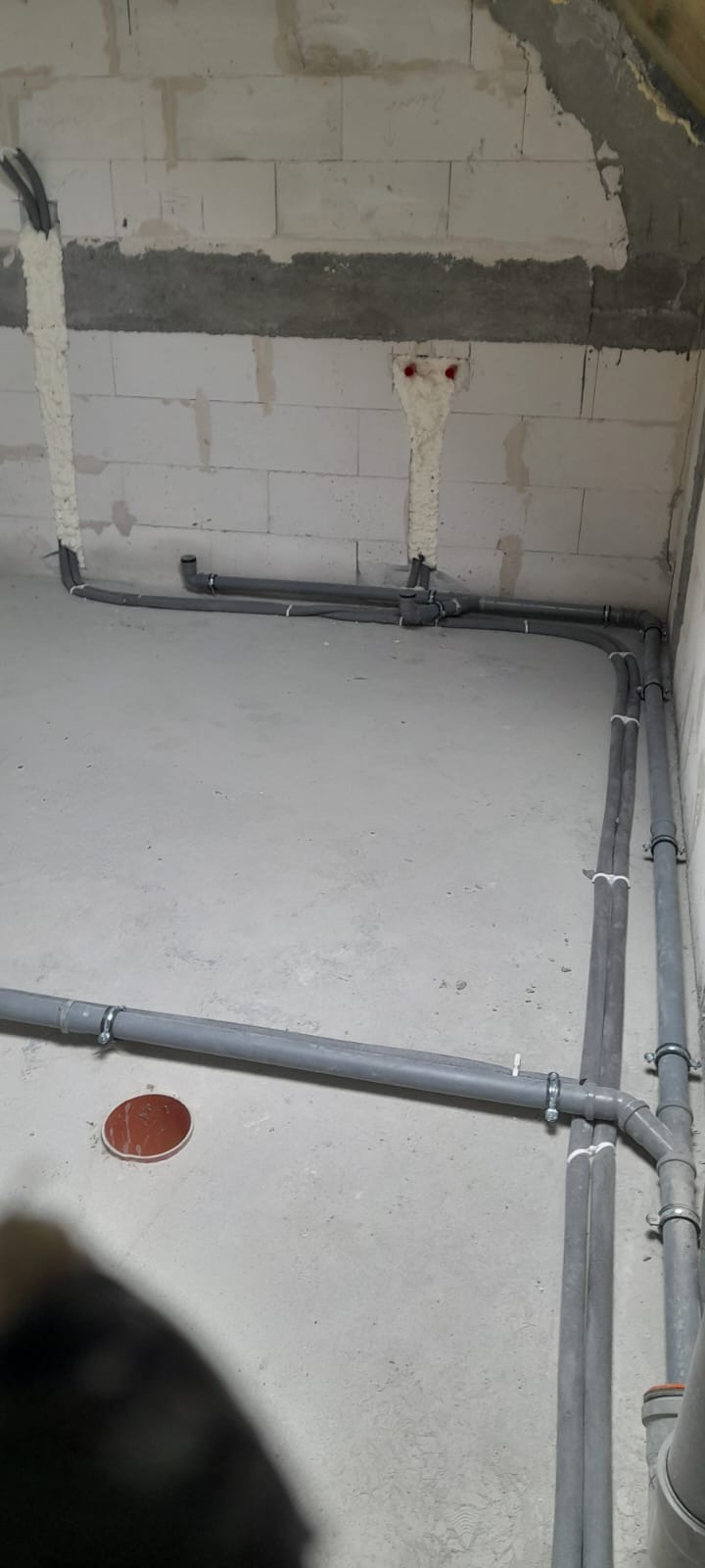 Szare rury instalacyjne prowadzone wzdłuż surowych ścian z betonu komórkowego, zabezpieczone pianką montażową w miejscach przejść, z widocznymi obejmami mocującymi i fragmentem podłogi z otworem.
