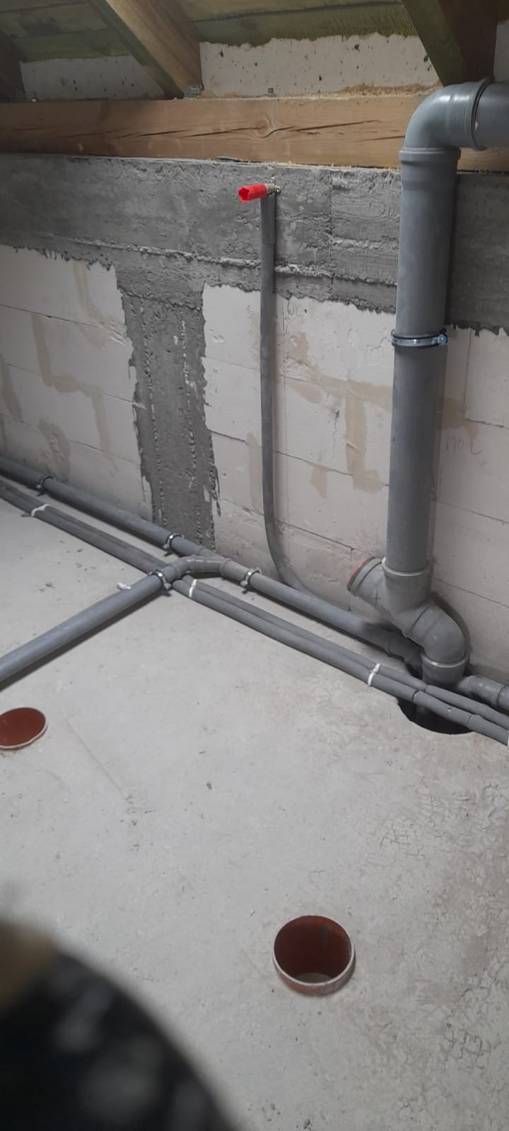 Szare rury instalacyjne prowadzone wzdłuż surowej ściany z betonu komórkowego i betonowej podłogi, z widocznymi otworami w podłodze i czerwoną rączką zaworu na pionowej rurze.