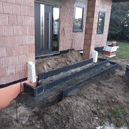 MR KOP - Budowa schodów wejściowych do domu z cegły. Widoczne szalunki i częściowo wypełnione betonem stopnie. W tle drzwi wejściowe i okna.