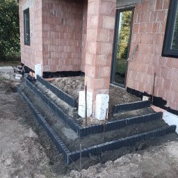 MR KOP - Budowa schodów zewnętrznych z betonowych elementów przy budynku z czerwonej cegły. Widoczne etapy prac, szare elementy konstrukcyjne i piasek.