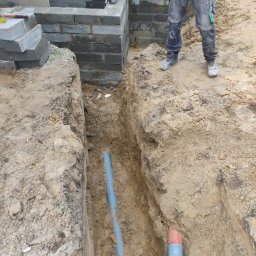 MR KOP - Wykopany wąski rów w ziemi z ułożonymi rurami, obok fundament z szarych bloczków. Widoczny fragment osoby w roboczych spodniach. Prace ziemne w trakcie realizacji.