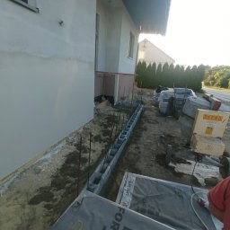 MR KOP - Budowa fundamentów: bloczki betonowe z wystającymi prętami zbrojeniowymi przy ścianie budynku. Na ziemi leżą materiały budowlane i stoi betoniarka.