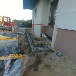 MR KOP - Fundament pod budynek w trakcie budowy: bloczki betonowe, zbrojenie, wykop. W tle koparka i materiały budowlane. Widoczne elementy elewacji budynku.