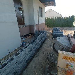MR KOP - Budowa murku z szarych bloczków betonowych przy domu jednorodzinnym. Widoczna betoniarka i zapas bloczków. Prace w toku, zbrojenie wystaje z bloczków.