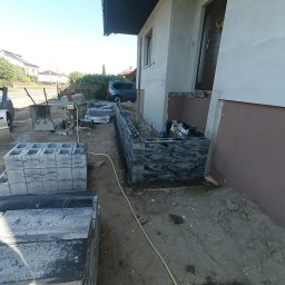 MR KOP - Budowa murku z bloczków betonowych przy domu jednorodzinnym. Widoczne materiały budowlane, narzędzia i częściowo wykonana konstrukcja. Słoneczny dzień.