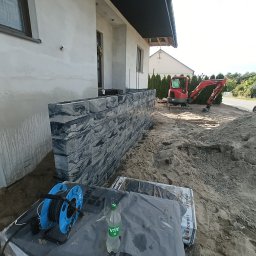 MR KOP - Wznoszenie murku z szarej kostki betonowej przy budynku. W tle minikoparka i teren w trakcie niwelacji. Na pierwszym planie narzędzia i materiały budowlane.