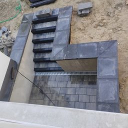 MR KOP - Ukończone schody kamienne z kostki brukowej w Płusach. Widok z góry na szare stopnie i obrzeża, z elementami drenażu liniowego i niezabudowanym terenem w tle.