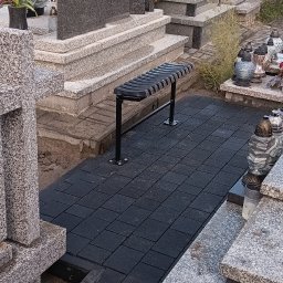 MR KOP - Ciemna kostka brukowa ułożona na cmentarzu między nagrobkami, z czarną ławką. Widok z góry, oświetlenie naturalne, nagrobki z kwiatami w tle.