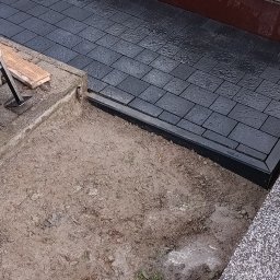 MR KOP - Częściowo ułożona, ciemna kostka brukowa wokół nagrobka z szarego granitu. Fragment wykopu z ziemią. Widoczne kwiaty i znicze na nagrobku.