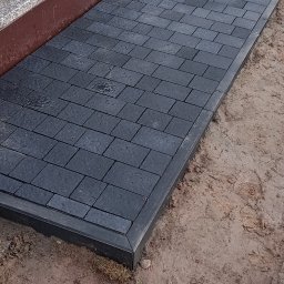 MR KOP - Ułożona, grafitowa kostka brukowa przy nagrobku, z widocznym obramowaniem. Świeżo ułożona nawierzchnia na cmentarzu, ziemia wokół.