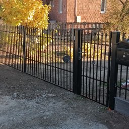 MR KOP - Czarny, metalowy płot z furtką i skrzynką na listy nr 24, widok z zewnątrz posesji. W tle dom z czerwonej cegły i drzewo z żółtymi liśćmi.