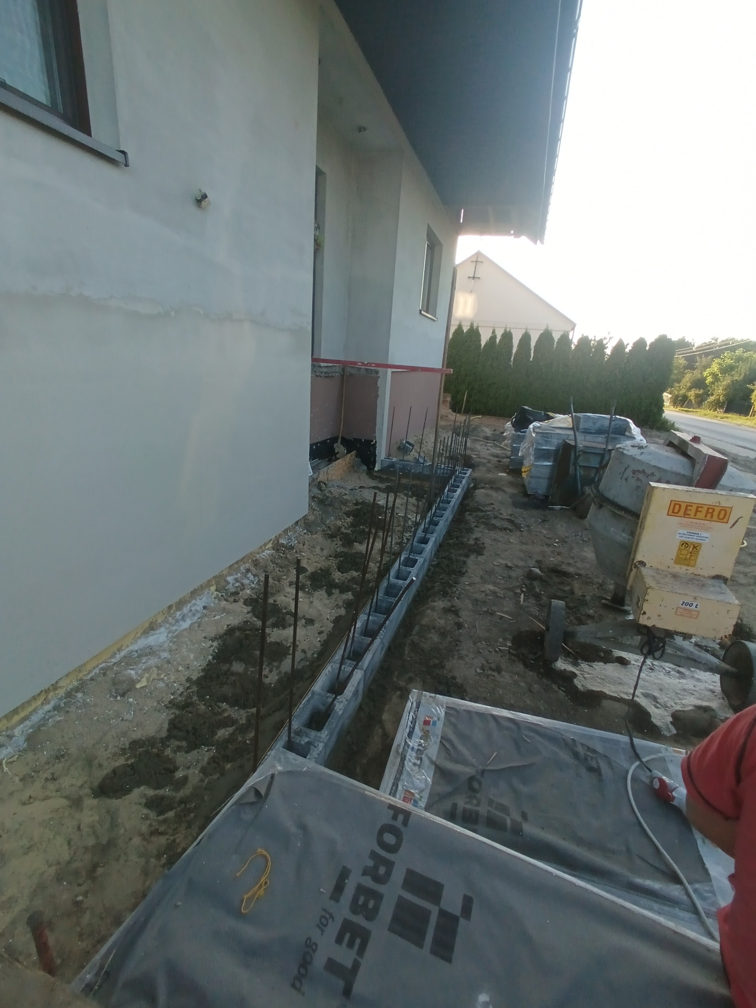 Budowa fundamentów: bloczki betonowe z wystającymi prętami zbrojeniowymi przy ścianie budynku. Na ziemi leżą materiały budowlane i stoi betoniarka.