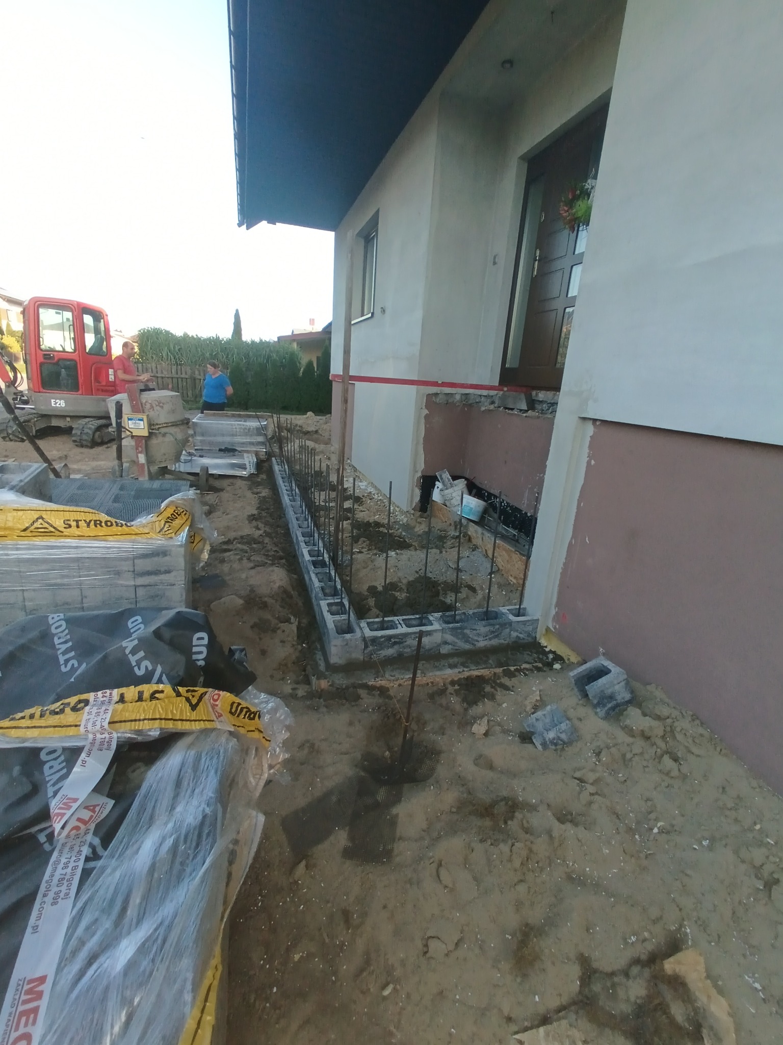 Fundament pod budynek w trakcie budowy: bloczki betonowe, zbrojenie, wykop. W tle koparka i materiały budowlane. Widoczne elementy elewacji budynku.