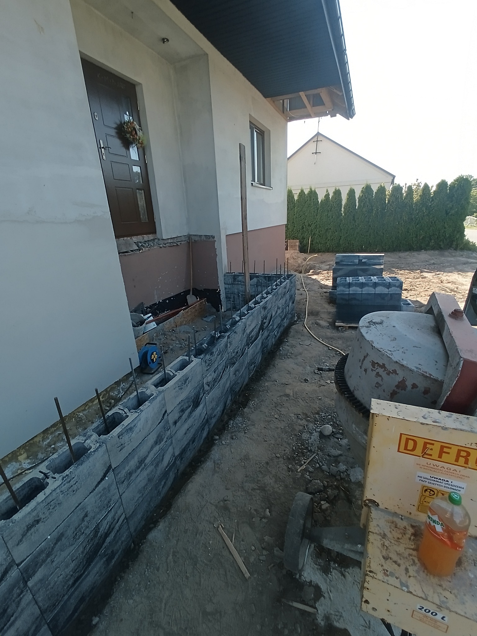 Budowa murku z szarych bloczków betonowych przy domu jednorodzinnym. Widoczna betoniarka i zapas bloczków. Prace w toku, zbrojenie wystaje z bloczków.
