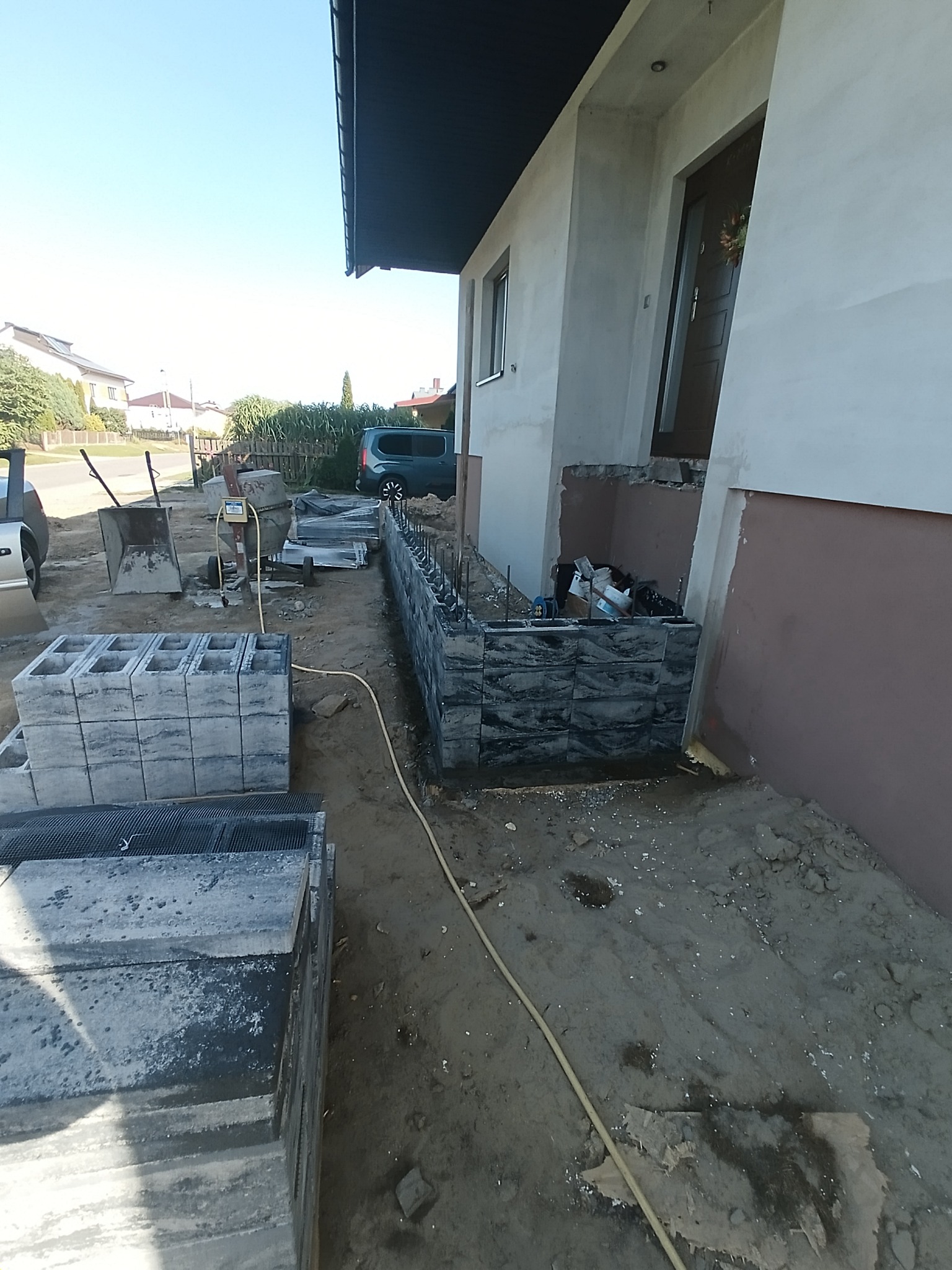 Budowa murku z bloczków betonowych przy domu jednorodzinnym. Widoczne materiały budowlane, narzędzia i częściowo wykonana konstrukcja. Słoneczny dzień.