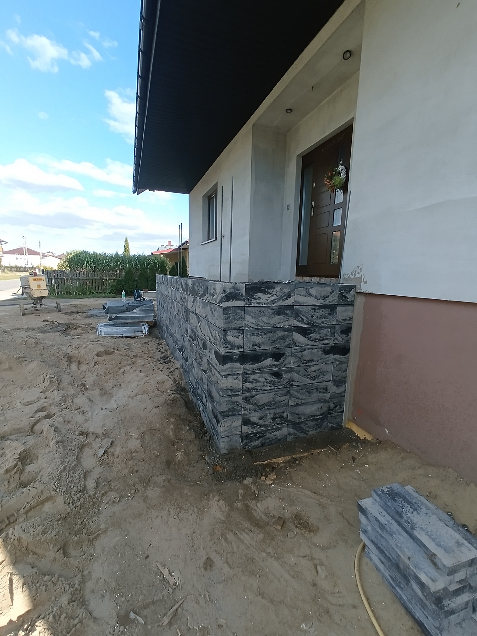 Wykończony cokół domu z szarej, wzorzystej kostki, widoczny z bliska, z elementami placu budowy w tle. Wejście do domu z dekoracyjnym wieńcem.