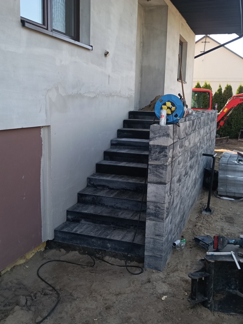 Zewnętrzne schody z ciemnej kostki, prowadzące do budynku z surowym tynkiem. Widoczny fragment ściany oporowej z bloczków i narzędzia budowlane. W tle koparka.