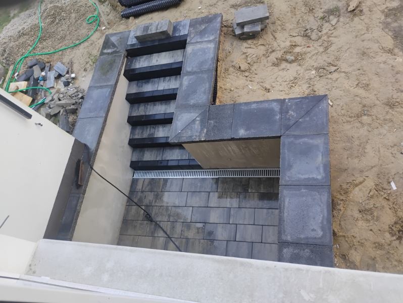 Ukończone schody kamienne z kostki brukowej w Płusach. Widok z góry na szare stopnie i obrzeża, z elementami drenażu liniowego i niezabudowanym terenem w tle.